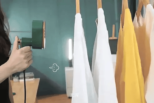 A person using a portable mini iron on clothes hanging on a rack against a green wall.